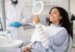 A woman sits in a dental chair with a bib, smiling at her reflection in a handheld mirror. A dentist in a white coat, mask, and blue gloves stands nearby in a bright, modern dental office.